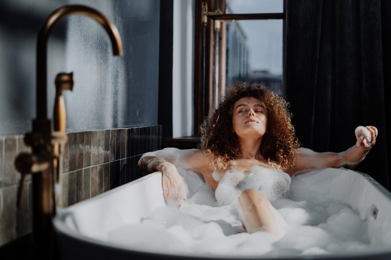 Young woman with curly hair relaxing in a sunlit bathtub filled with bubbles, enjoying a serene bath time.
