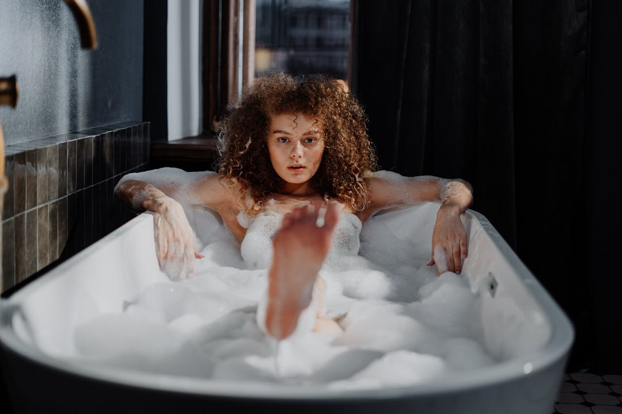 Young woman with curly hair relaxing in a bathtub filled with soap bubbles.